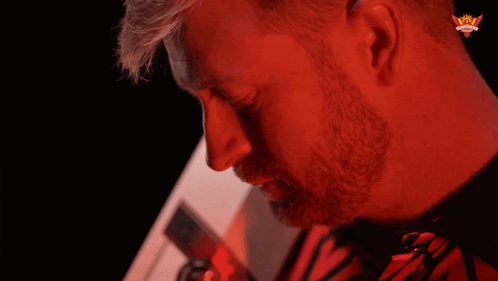 A Man In A Red Shirt With A Crown On His Ear Looks At The Camera GIF