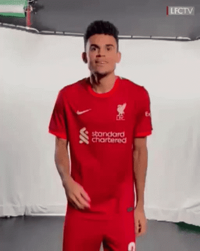 A Man In A Red Soccer Uniform Is Standing In Front Of A White Wall And Smiling . GIF