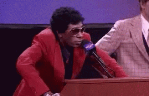 A Man In A Red Suit And Sunglasses Is Standing At A Podium . GIF