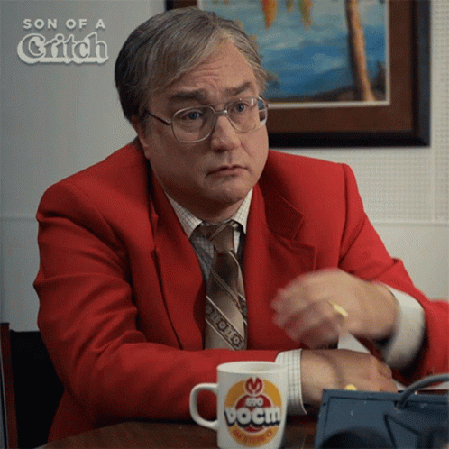 A Man In A Red Suit And Tie Is Sitting At A Table With A Cup That Says Boom GIF