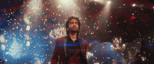 A Man In A Red Suit Stands On A Stage With Confetti Falling Around Him And A Sign That Says Peace Makes Love Stronger GIF