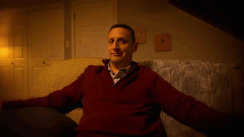 A Man In A Red Sweater And Tie Is Sitting On A Couch GIF
