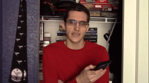 A Man In A Red Sweater Is Holding A Cell Phone In Front Of A Shelf With A Board Game That Says Transformers GIF