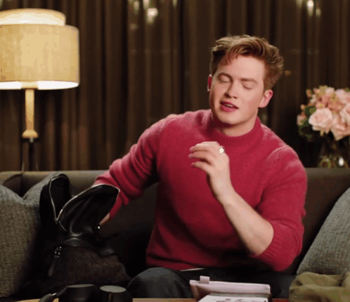 A Man In A Red Sweater Sits On A Couch Holding A Black Bag GIF