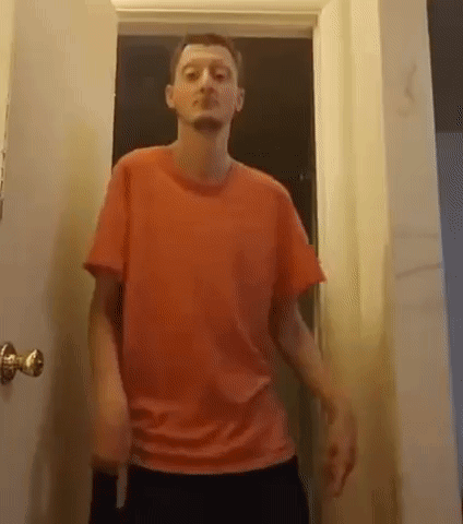A Man In A Red T-Shirt Is Standing In A Doorway . GIF