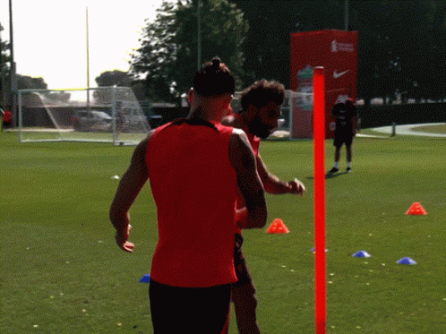 A Man In A Red Tank Top Is Standing Next To Another Man GIF
