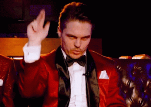 A Man In A Red Tuxedo And Bow Tie Is Sitting On A Couch Making A Gesture With His Hand . GIF