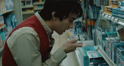 A Man In A Red Vest Is Looking At A Box Of Cold Medicine In A Store GIF