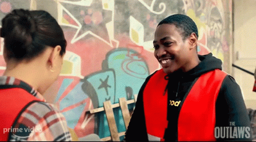 A Man In A Red Vest Is Smiling While Talking To A Woman In Front Of A Graffiti Wall . GIF