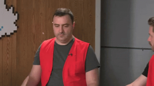 A Man In A Red Vest Is Standing Next To Another Man In A Gray Shirt . GIF