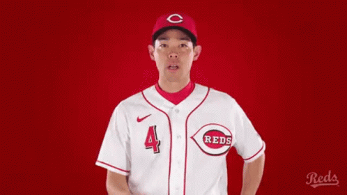 A Man In A Reds Baseball Uniform Is Pointing At Something . GIF