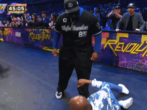 A Man In A Revolution 85 Jersey Stands Next To A Man Laying On The Ground GIF