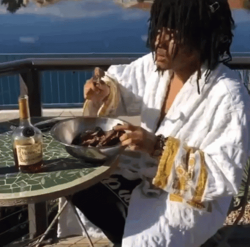 A Man In A Robe Is Sitting At A Table With A Bottle Of Hennessy GIF