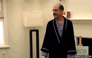 A Man In A Robe Is Standing In A Living Room With A Lamp . GIF