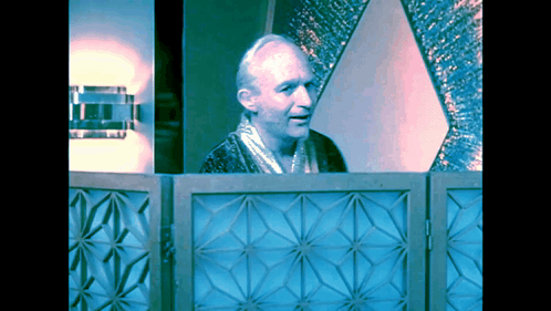 A Man In A Robe Stands Behind A Blue Geometric Screen GIF