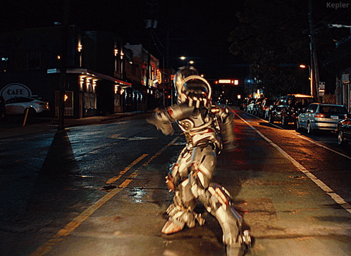 A Man In A Robot Costume Is Walking Down A Street At Night In Front Of A Cafe Sign GIF
