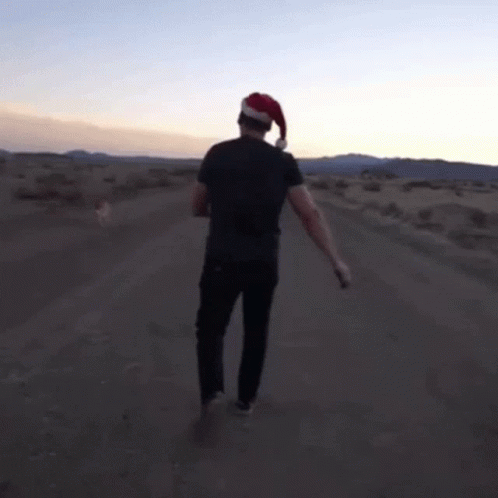A Man In A Santa Hat Is Walking Down A Dirt Road GIF