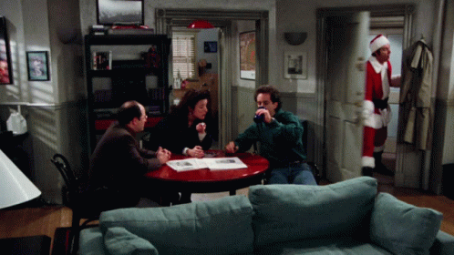 A Man In A Santa Suit Stands In A Living Room With A Group Of People Sitting Around A Table GIF