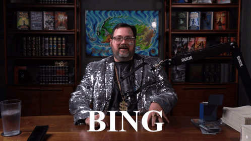 A Man In A Sequined Jacket Is Sitting At A Desk With The Word Bing Written On It GIF
