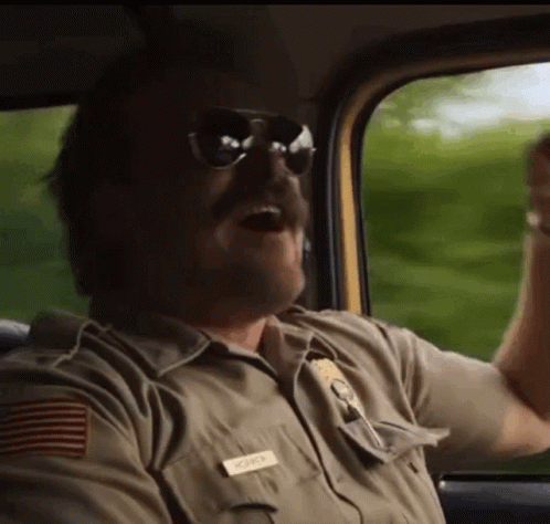 A Man In A Sheriff 'S Uniform And Sunglasses Is Driving A Car . GIF