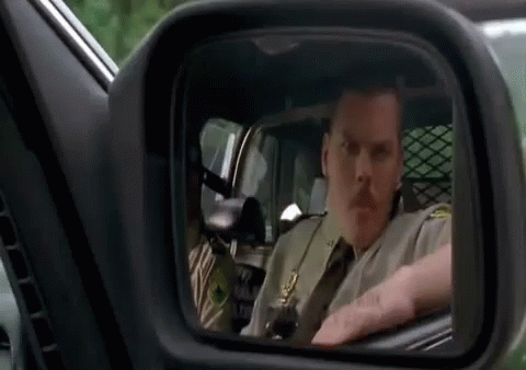 A Man In A Sheriff 'S Uniform Is Driving A Car And Looking In The Rear View Mirror . GIF