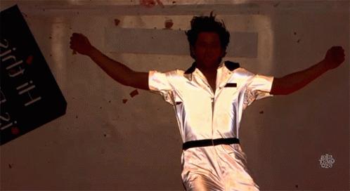 A Man In A Silver Jumpsuit Is Dancing In Front Of A Sign That Says ' Bitch ' GIF