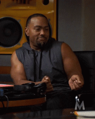 A Man In A Sleeveless Hoodie Is Sitting In Front Of A Masterclass Logo GIF