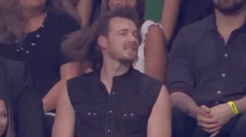 A Man In A Sleeveless Shirt Is Sitting In A Crowd GIF