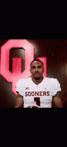 A Man In A Sooners Jersey Is Holding A Football In Front Of A Large Red Q . GIF