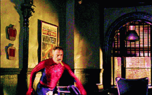A Man In A Spiderman Costume Is Standing In A Room Next To A Sign That Says Wanted . GIF