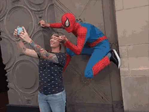 A Man In A Spiderman Costume Is Taking A Picture Of A Woman In A Tattooed Shirt . GIF