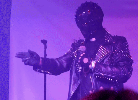 A Man In A Spiked Leather Jacket Is Singing Into A Microphone In A Dark Room GIF