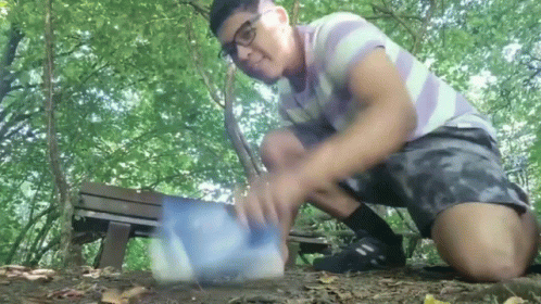 A Man In A Striped Shirt And Shorts Is Kneeling Down In The Woods Playing With A Ball GIF