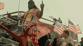 A Man In A Striped Shirt Is Giving The Middle Finger While Standing In Front Of An American Flag . GIF