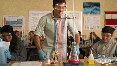 A Man In A Striped Shirt Is Standing In Front Of A Beaker With Red Liquid In It GIF