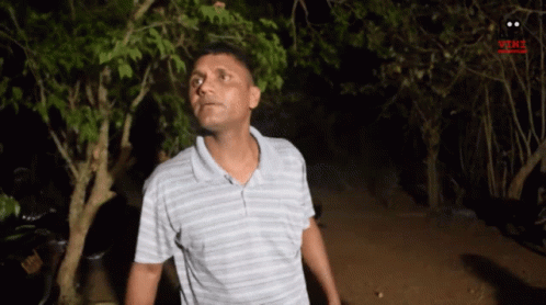 A Man In A Striped Shirt Is Standing In Front Of Trees At Night . GIF