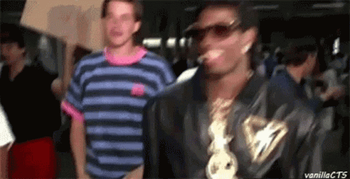 A Man In A Striped Shirt Is Standing Next To A Man Wearing Sunglasses And A Chain Around His Neck . GIF