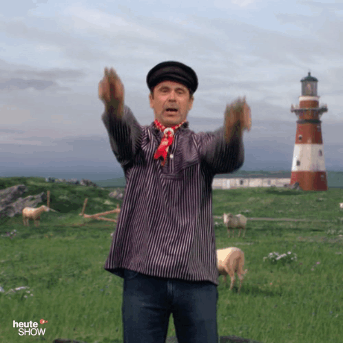 A Man In A Striped Shirt Stands In A Field With A Lighthouse In The Background And The Word Heute Show On The Bottom GIF