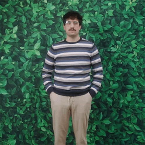 A Man In A Striped Sweater Stands With His Hands In His Pockets In Front Of A Green Wall GIF