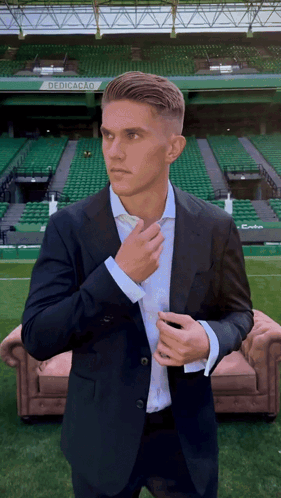 A Man In A Suit Adjusts His Tie In Front Of A Stadium That Says Dedicacao On It GIF