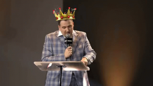 A Man In A Suit And A Crown Is Standing At A Podium With A Microphone . GIF