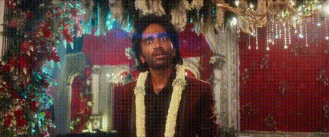A Man In A Suit And A Garland Around His Neck Is Standing In Front Of A Red Curtain . GIF
