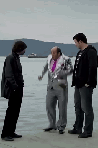 A Man In A Suit And A Purple Tie Talks To Two Other Men In Front Of A Body Of Water GIF
