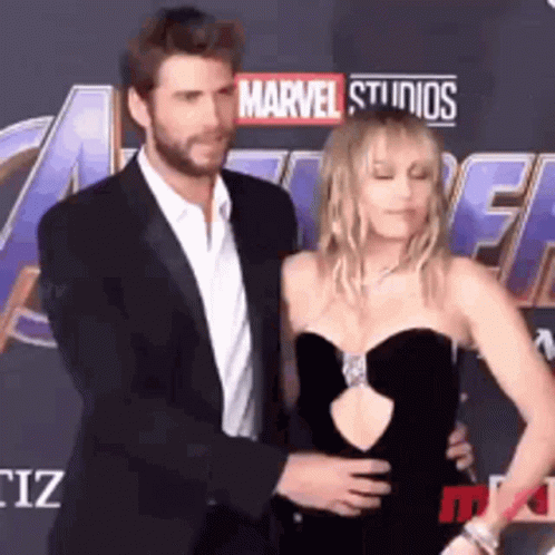 A Man In A Suit And A Woman In A Black Dress Are Standing On A Red Carpet . GIF