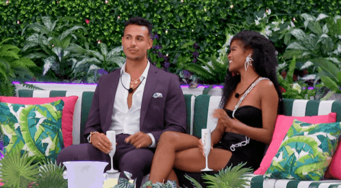 A Man In A Suit And A Woman In A Black Dress Sit On A Couch GIF