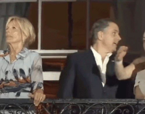A Man In A Suit And A Woman In A Floral Shirt Are Standing On A Balcony GIF