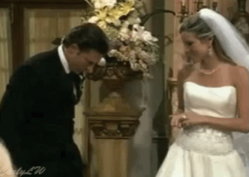 A Man In A Suit And A Woman In A Wedding Dress Are Standing Next To Each Other In Front Of A Vase Of Flowers GIF