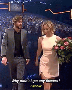 A Man In A Suit And A Woman In A White Dress Holding A Bouquet Of Flowers Are Walking On A Stage . GIF