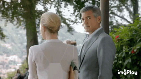 A Man In A Suit And A Woman In A White Shirt Are Looking At Each Other . GIF