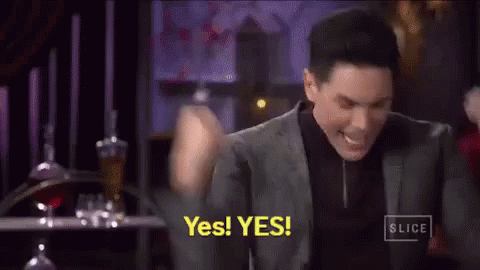 A Man In A Suit And Black Shirt Is Holding His Fist In The Air And Saying Yes . GIF